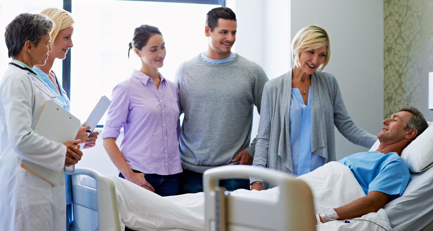 Patient with hospital indemnity insurance is laying in a hospital bed with family standing next to him.