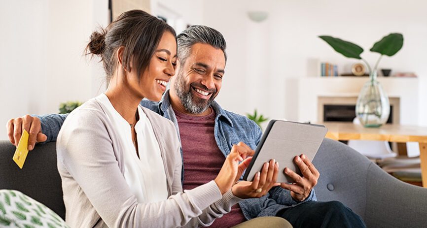 A couple is happily browsing on a tablet. A man is holding his credit card loosely, implying he is making a purchase.