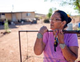 A Navajo woman standing by the gate of her home, with a concerned look on her face.