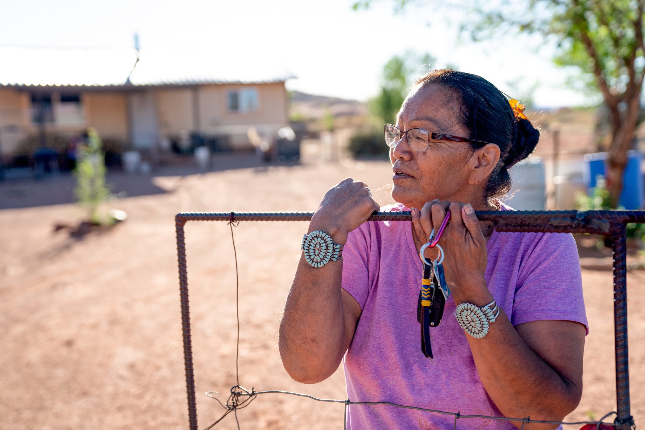 A Navajo woman standing by the gate of her home, with a concerned look on her face.