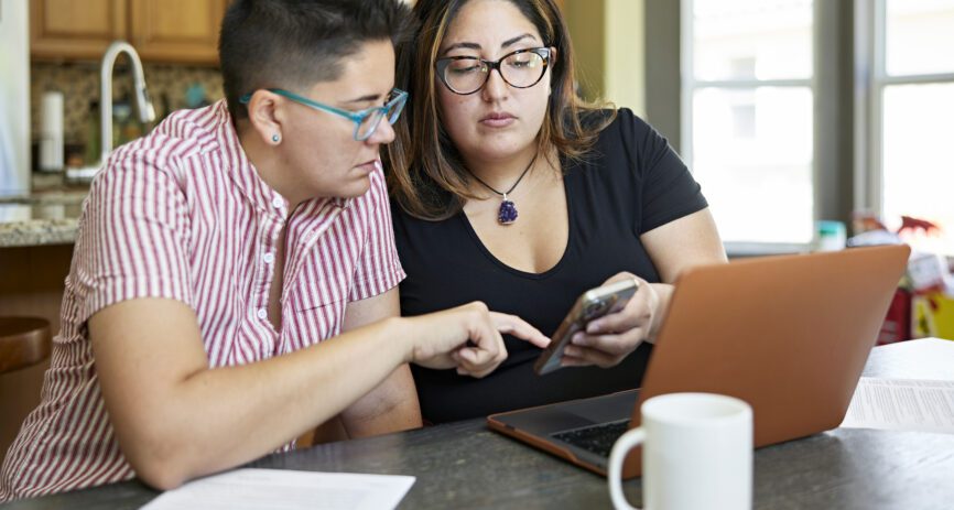 Two women wearing glasses look at a laptop and smartphone at their kitchen counter.