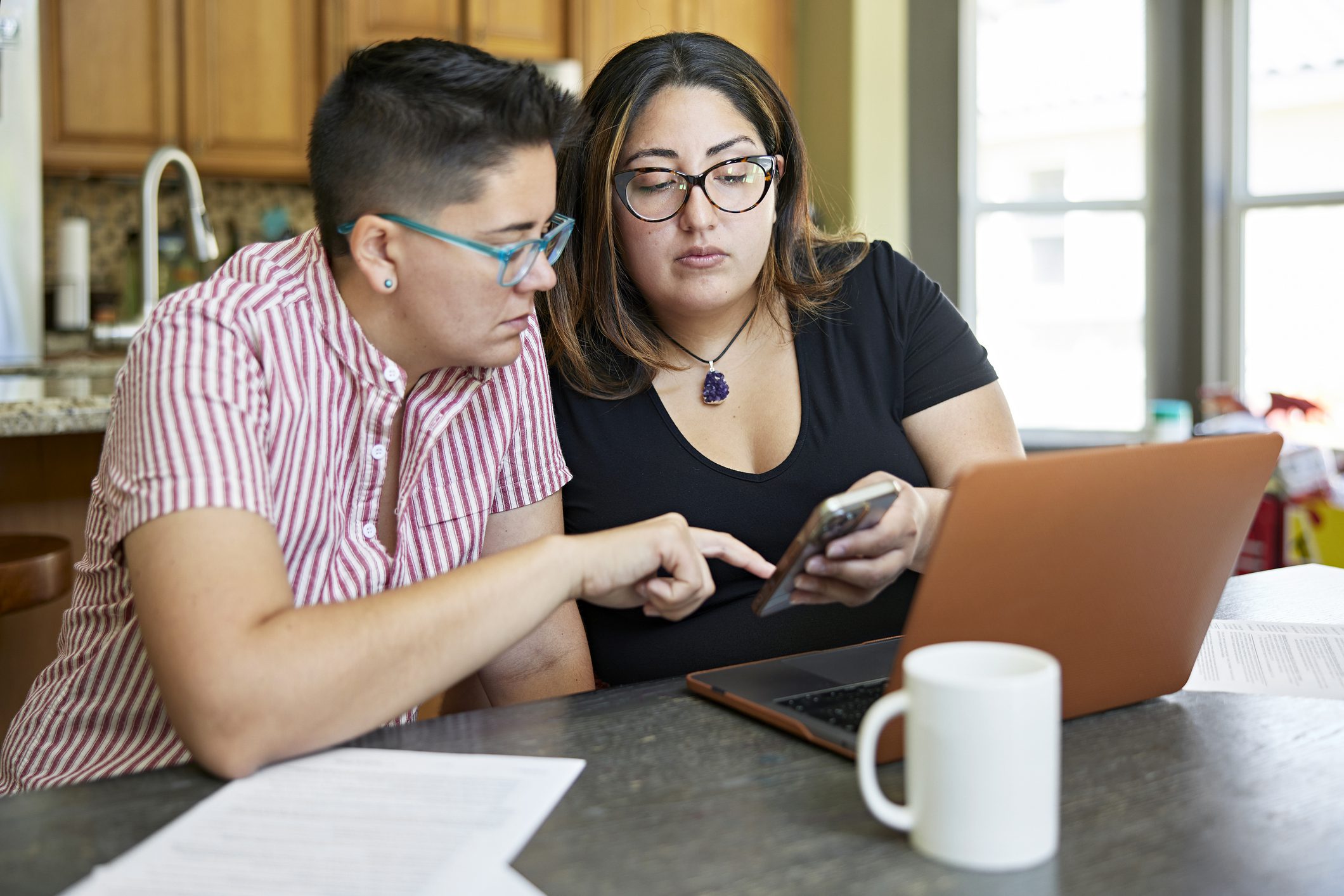 Two women wearing glasses look at a laptop and smartphone at their kitchen counter.