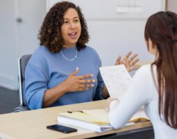 Woman HR manager talking across desk to woman employee holding a paper whose back is to camera.