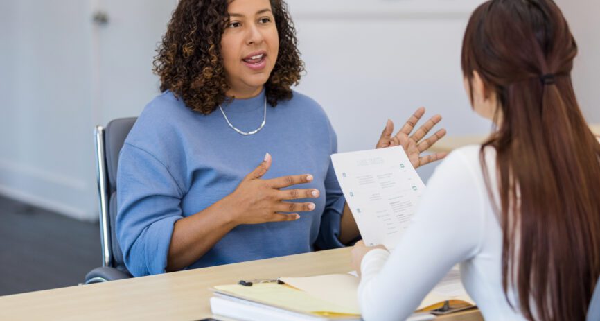 Woman HR manager talking across desk to woman employee holding a paper whose back is to camera.