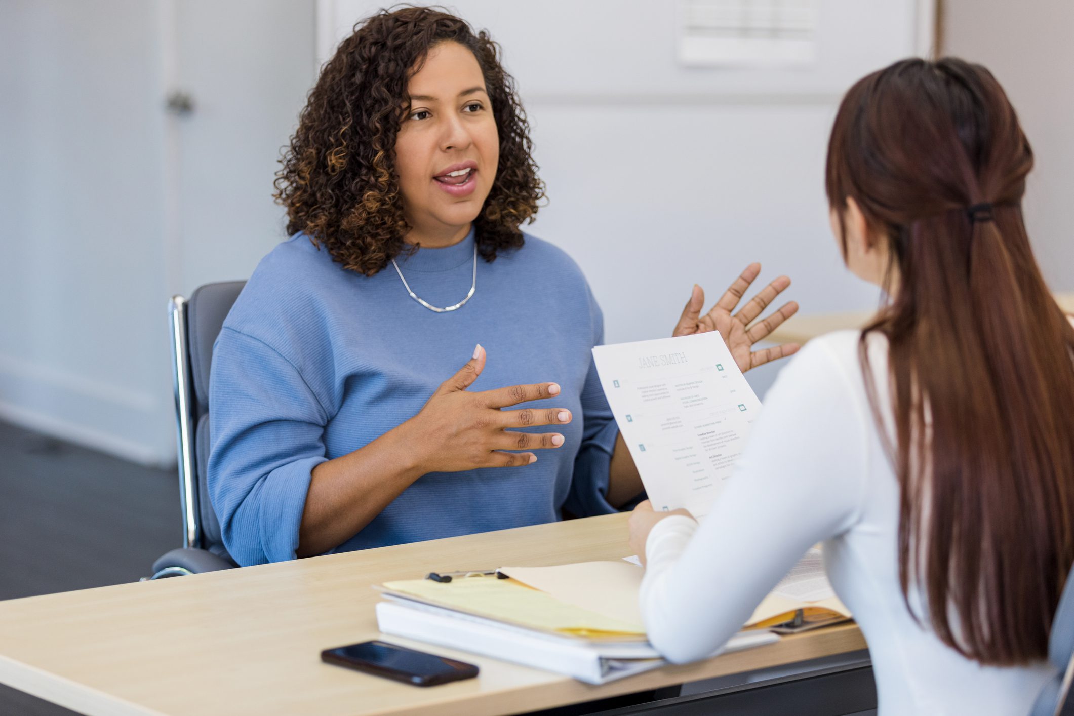 Woman HR manager talking across desk to woman employee holding a paper whose back is to camera.