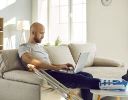 A man with a broken leg sits on his couch and pays a medial bill on his laptop.