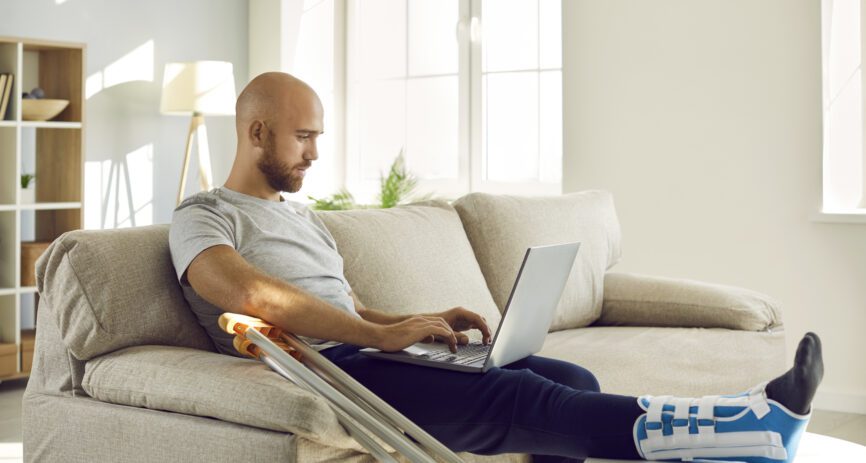 A man with a broken leg sits on his couch and pays a medial bill on his laptop.