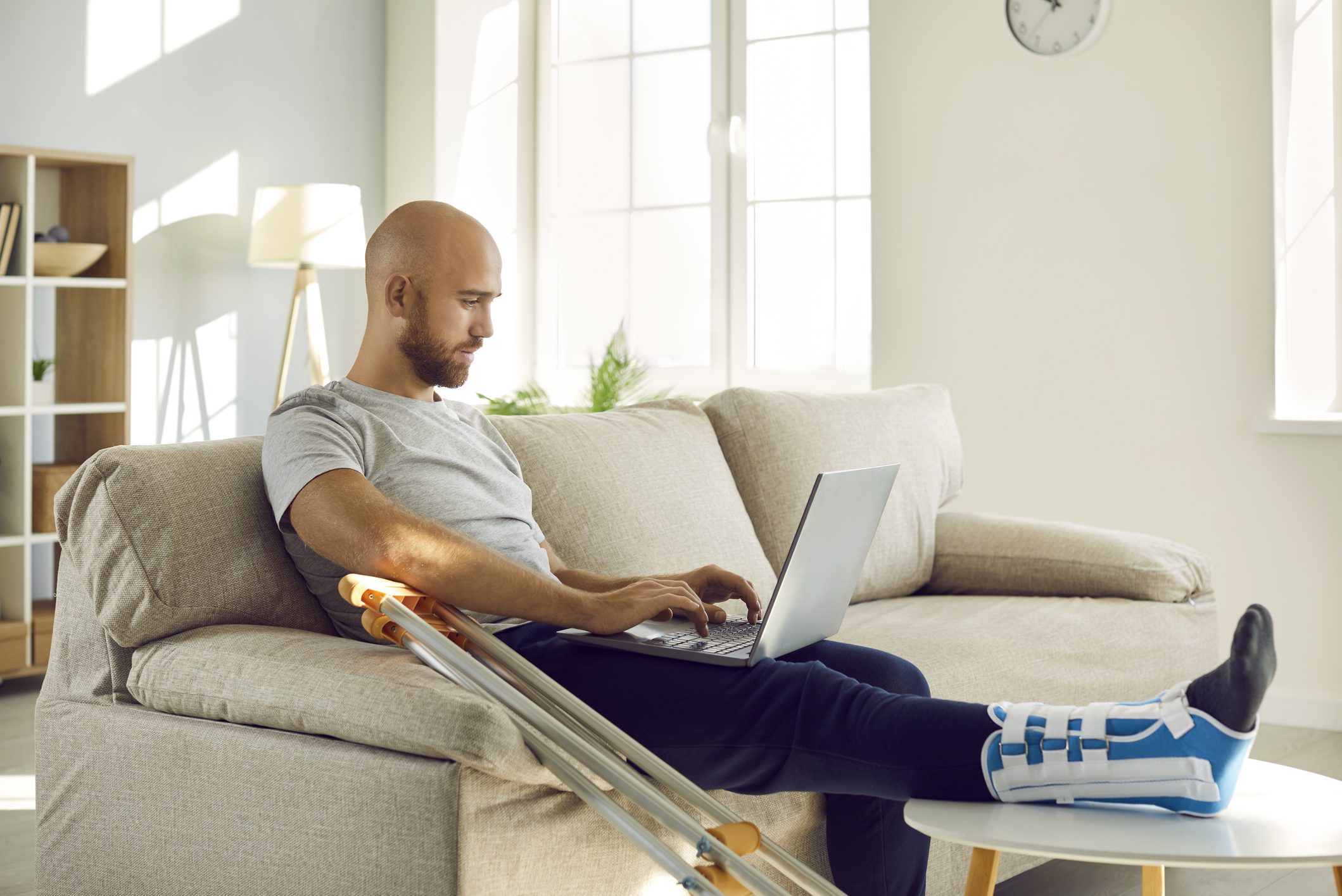 A man with a broken leg sits on his couch and pays a medial bill on his laptop.